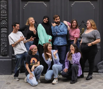 Groupe de neuf personnes posant de manière joyeuse et décontractée devant une façade sombre, illustrant l’ambiance dynamique et conviviale de l’équipe.