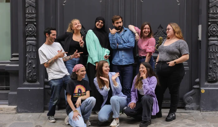 Groupe de neuf personnes posant de manière joyeuse et décontractée devant une façade sombre, illustrant l’ambiance dynamique et conviviale de l’équipe.