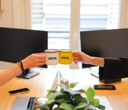 Cheers à l’esprit d’équipe chez HKM Deux personnes trinquent avec des mugs HKM dans un bureau lumineux, symbolisant la convivialité et l’esprit d’équipe de l’agence.