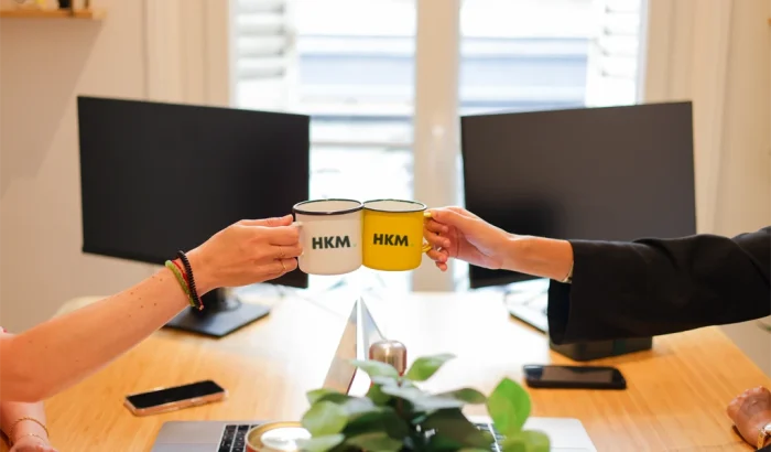 Deux personnes trinquent avec des mugs HKM dans un bureau lumineux, symbolisant la convivialité et l’esprit d’équipe de l’agence.